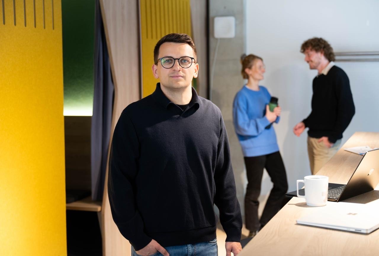 Ein Mann steht an einem Schreibtisch, schaut die Kamera an und zwei Menschen unterhalten sich im Hintergrund.